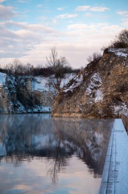 Sakin bir gölü olan sakin bir kış manzarası karlı uçurumları ve bulutlu bir gökyüzünün altındaki çıplak ağaçları yansıtıyor..