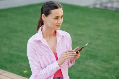 Pembe gömlekli hoş esmer kadın güneşli bir günde dışarıda akıllı telefon kullanıyor..