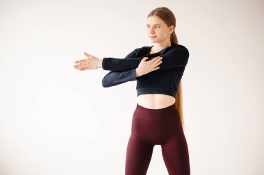 Uzun sarı saçlı fit bir genç kadın, minimalist bir stüdyo ortamında atletik kıyafetler giyer ve omuz gerer.