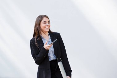 Takım elbiseli neşeli, profesyonel bir kadın elinde dizüstü bilgisayar ve akıllı telefonuyla yürüyüp bir sohbete girişiyor.