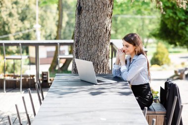 Gülümseyen bir iş kadını, doğayla çevrili açık bir kafe masasında dizüstü bilgisayarıyla çalışarak verimli bir günün tadını çıkarıyor.