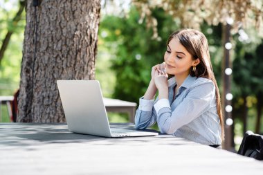 Odaklanmış genç bir kadın, yemyeşil ve benekli güneş ışığının ortasında açık bir kafe masasında otururken bilgisayarında verimli bir çalışma seansının keyfini çıkarıyor.