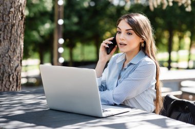 Profesyonel bir kadın, yeşil ve benekli güneş ışığıyla çevrili, açık bir kafede bilgisayarla telefon görüşmesi yapıyor.