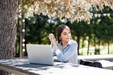 Mavi çizgili tişörtlü odaklanmış genç bir kadın bir dizüstü bilgisayarla ahşap bir masada oturuyor, bir kafe ortamının doğal ışık ve açık hava ortamının tadını çıkarıyor.