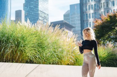 Formda, spor kıyafetleri içinde sarışın bir kadın şehir yeşili ve modern mimarinin ortasındaki akıllı telefonuna bakmak için yürüyüşünü durduruyor.