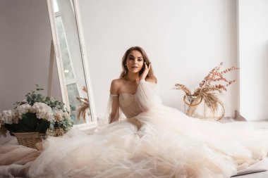 Parlak bir stüdyo ortamında geniş bir ayna ve çiçekli bir elbise içindeki güzel bir gelin ya da manken zarif bir şekilde oturur.