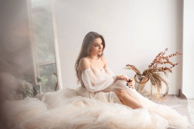 Uzun kahverengi saçlı, güzel bir genç kadın omuzları açık ruhani bir elbise giyiyor, etrafında geniş bir tül etekle oturuyor, düşünceli bir şekilde yan tarafa bakıyor.