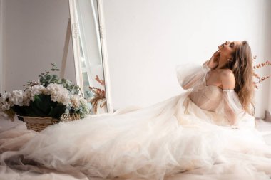 Zarif bir çiçek aranjmanı ve yumuşak kumaşın arasına oturmuş, şehvetli, omuzları açık bir elbise içinde sakin bir güzelliği yansıtan sakin bir gelin.