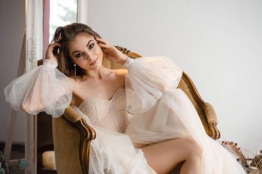 Dramatik makyajlı güzel bir kadın, eski bir sandalyede dinlenirken, kolları kaslı, narin, omuz dışı bir gecelik giyer.