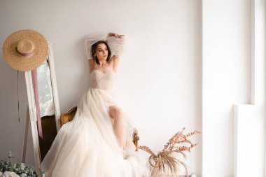 Fildişi bir gelinlik giymiş genç bir kadın şehvetli kolları olan zarif bir şekilde oturuyor, kolları havada, parlak, havalı bir stüdyo ortamında bohem dekor unsurlarıyla.