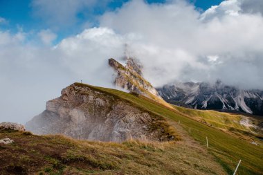 İtalyan Dolomitleri 'nde, Seceda' nın çimenli sırtında tek başına yürüyen bir yürüyüşçü, sarp dağ tepelerinin etrafında dönen dramatik bulutlarla...