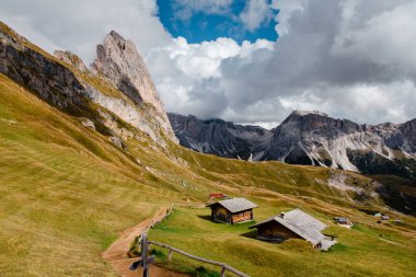 Dolomitlerin ikonik sivri tepeleri, dinamik bir gökyüzünün altında ahşap kiremitlerle benekli yuvarlanan yeşil tepelerin üzerinde yükselir.