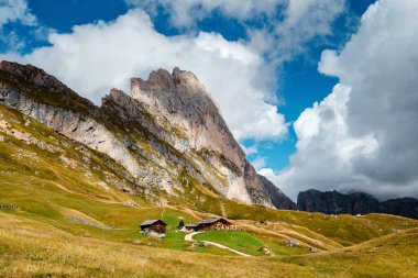 İtalyan Dolomitlerinin Seceda tepesinin manzarası. Dinamik bir gökyüzünün altında yeşil yuvarlanan tepelere yerleşmiş ahşap kiremitler.