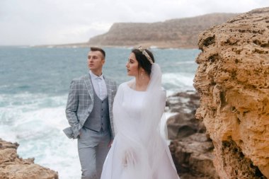 Yeni evli bir çift engebeli bir kıyı şeridinde duruyor, düğün kıyafetleri doğal, vahşi çevre ve çalkantılı deniz ile çelişiyor.