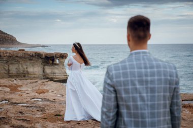 Beyaz elbiseli bir gelin ve takım elbiseli bir damat Kıbrıs 'taki Akdeniz manzaralı kayalık kıyılarda duruyorlar.