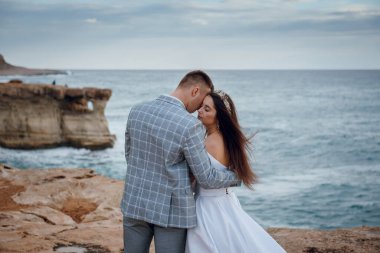 Gelin ve damat, arka planda okyanus dalgalarının çarptığı kayalık bir sahil şeridinde şefkatle kucaklaşır.