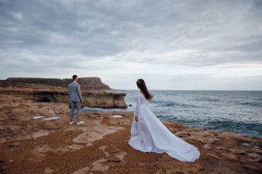 Dalgalı beyaz elbiseli bir gelin ve takım elbiseli bir damat Kıbrıs 'ta denize bakan kayalık bir kıyı şeridinde duruyorlar.