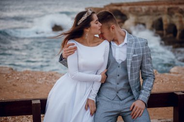 Yeni evli bir çift, arka planda dalgaların çarptığı kayalık bir sahil şeridinde şefkatle kucaklaşır.