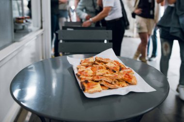 Arka planda insanların olduğu bir etkinlikte taze kesilmiş pizza dilimleri servis edilmeye hazır.