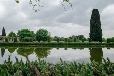 Huzurlu bir yansıma göleti, bulutlu bir gökyüzünün altında tarihi bir İtalyan bahçesinde manikürlü çalılar ve selvi ağaçları yansıtır.