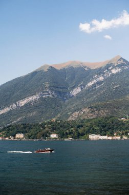 Como Gölü 'nün derin mavi sularını hızla geçen bir motorlu tekne kıyı şeridinin arkasında yükselen yemyeşil dağlarla berrak bir gökyüzünün altında köyler.