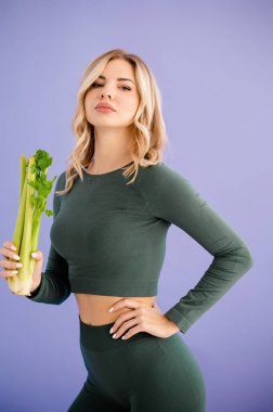 Atletik giyinen fit bir kadın, sağlıklı beslenme ve sağlığını sembolize eden sağlam bir mor arka plana karşı bir demet taze kereviz sapıyla güven içinde poz verir.