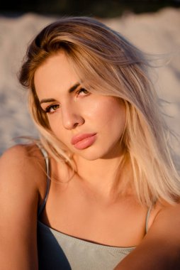 Sarışın ve çarpıcı gözlü genç bir kadın kumlu bir sahilde batan güneşin sıcacık parıltısıyla aydınlanır.