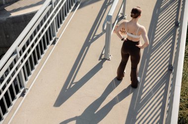 Spor sutyenli ve taytlı bir kadının arka görünüşü yaya köprüsünde duruyor, elleri kalçasında, açık havada eğleniyor.