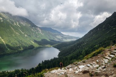 Yalnız bir yürüyüşçü bulutlu bir gökyüzünün altında yemyeşil dağlarla çevrili sakin Morskie Oko Gölü 'ne bakan kayalık bir yola yükselir.