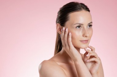 Parlak ciltli genç bir kadın parmak uçlarıyla yüzüne dokunuyor, doğal güzelliğini ve cildini ön plana çıkarıyor.