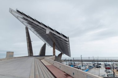 Çarpıcı bir modern güneş enerjisi tesisi bulutlu bir gökyüzünün altında teknelerle dolu hareketli bir limanın yanında belirgin bir şekilde duruyor.