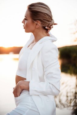 Beyaz ceket giyen şık bir kadın ve altın saat boyunca pantolonu gölün kenarında duruyor, eli hafifçe topuzuna dokunuyor.