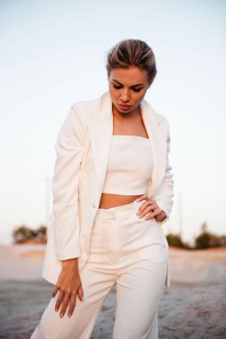 Beyaz bir takım elbise giyen şık bir kadın güneş batarken açık havada güvenli bir şekilde duruyor ve sıcak bir parıltı yaratıyor.