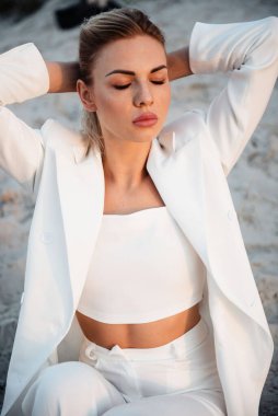 Şık beyaz takım elbiseli sakin bir kadın kumun üzerinde oturuyor gözleri kapalı ve elleri başının arkasında, ılık güneş ışığıyla yıkanmış.