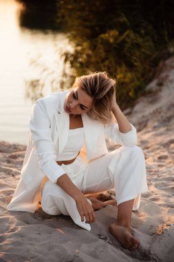 Beyaz takım elbiseli şık bir kadın, güneş batarken kumlu bir sahilde poz veriyor. Sudaki altın ışığı ve kıyafetlerini yansıtıyor.