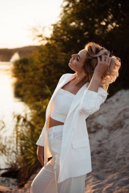 Beyaz takım elbiseli şık bir kadın ve açık havada poz veriyor güzel bir gün batımında, saçları rüzgarla dalgalanırken.