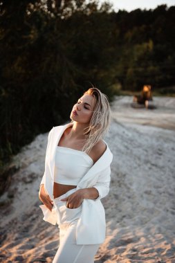 Şık, sarı saçlı bir kadın gün batımının sıcacık parıltısıyla güneşleniyor sakin bir plajda, huzurlu bir tefekkür içinde gözleri kapalı.