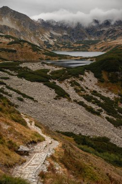 Dolambaçlı bir taş yol engebeli bir dağlık araziye çıkar dağınık yeşil çalılar ve bulutlu bir gökyüzünün altındaki sakin bir dağ gölüne doğru geniş bir yamaç.
