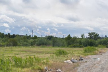 Doğal bir manzaranın geniş bir görüntüsü: yeşil bir tarla ve bulutlu bir gökyüzünün altındaki ağaçlarla kaplı tepeler. Ufukta bir sanayi fabrikasının sigara içme boruları görünüyor ve dramatik bir ekolojik kontrastı yaratıyor..