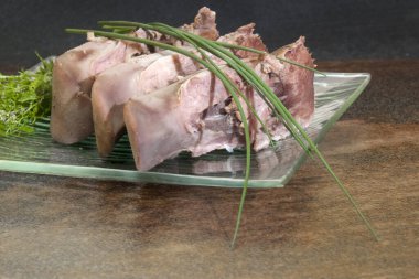 Üç büyük porsiyon kaynamış domuz dili, soğan ve dereotuyla süslenmiş, desenli cam bir tabakta servis ediliyor. Düşük açı ve karanlık arka plan aşçılık lüksü hissi yaratıyor..