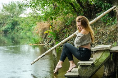 Genç güzel kadın modeli uzun sarı saçlı oturan farklı duygular kahkaha, üzüntü, keder, dalgınlık Nehri ve su almak için ormanın kıyısında ahşap bir köprü ile