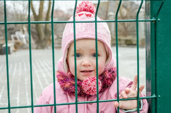 Lilla vackra flicka baby gråter med tårar i ögonen och en sorgliga, sorgliga känslor stängs ned som ett straff för ett staket av metall galler vill ha frihet, hatt och dunjacka — Stockfoto