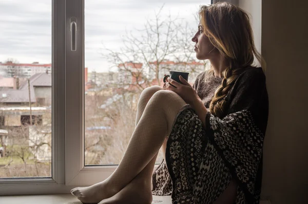 年轻漂亮的女孩学生坐在窗台上在窗口俯瞰全城和喝一杯热咖啡 — 图库照片