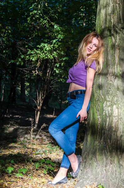 Joven hermosa chica sexy modelo rubio con pelo largo y rubio en jeans y chaqueta posando en el bosque entre los árboles y la vegetación —  Fotos de Stock