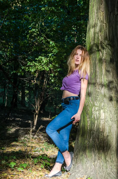 Joven hermosa chica sexy modelo rubio con pelo largo y rubio en jeans y chaqueta posando en el bosque entre los árboles y la vegetación —  Fotos de Stock