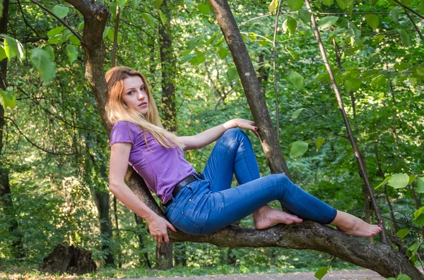 Joven hermosa chica sexy modelo rubio con pelo largo y rubio en jeans y chaqueta posando en el bosque entre los árboles y la vegetación —  Fotos de Stock