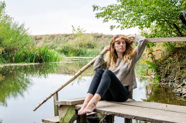 Jonge mooie vrouw model met lang blond haar zitten met verschillende emoties gelach, droefheid, verdriet, bedachtzaamheid op een houten brug aan de oever van de rivier en het bos op zoek naar water — Stockfoto