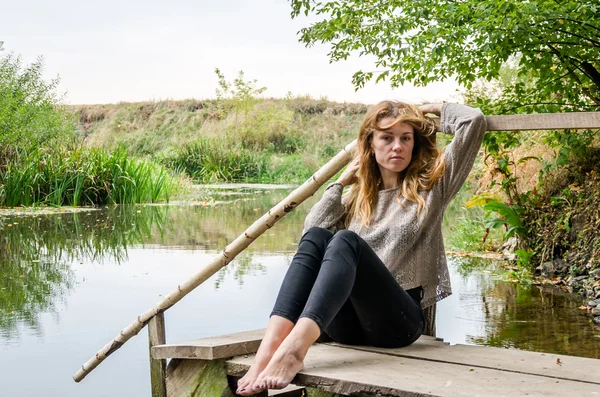 Joven modelo hermosa mujer con el pelo largo y rubio sentado con diferentes emociones risa, tristeza, dolor, consideración en un puente de madera en la orilla del río y el bosque en busca de agua — Foto de Stock