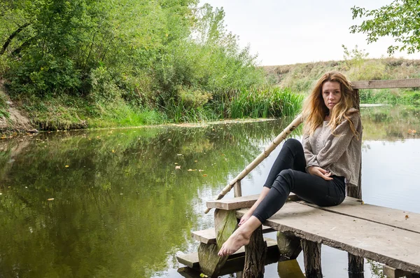 Joven modelo hermosa mujer con el pelo largo y rubio sentado con diferentes emociones risa, tristeza, dolor, consideración en un puente de madera en la orilla del río y el bosque en busca de agua — Foto de Stock