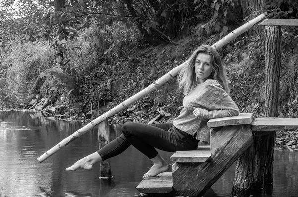 Giovane bella modella donna con lunghi capelli biondi seduta con diverse emozioni risate, tristezza, dolore, premura su un ponte di legno sulla riva del fiume e la foresta in cerca di acqua — Foto Stock